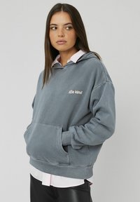 Hoodie oversize gris avec une poche kangourou, arborant "Soyez gentil" brodé en rose clair. Porté par-dessus une chemise rayée rose.