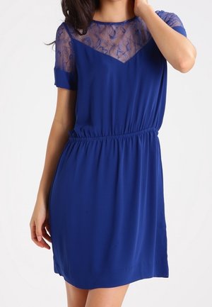 Femme portant une robe courte bleu roi avec des détails en dentelle sur le haut de la poitrine et les manches, taille élastique, debout sur un fond blanc.