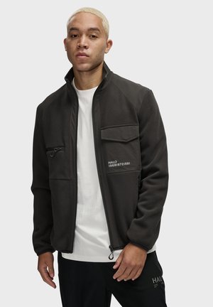 Ung mand med kort blond hår iført en sort zip-up jakke over en hvid skjorte, stående foran en ensfarvet baggrund.