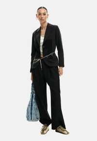 Blazer nero a righe sottili, top bianco, pantaloni neri a gamba larga, cintura a catena con cuore metallico, borsa in denim blu, sneakers marroni.