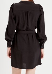 Robe noire à manches bouffantes, taille ceinturée, détail plié dans le dos et poignets boutonnés. Confectionnée dans un tissu texturé et léger.