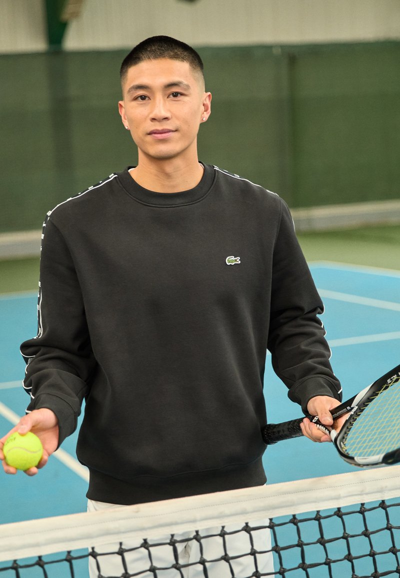 Schwarzer Sweatshirt mit seitlichen Streifen, mit kleinem grünen Krokodil-Logo. Hält einen grünen Tennisball und einen Schläger, in sportlicher Umgebung.
