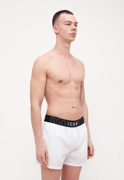 Shorts de bain blancs avec une taille élastique noire portant le texte "ICON" en blanc. Le tissu semble léger et lisse.