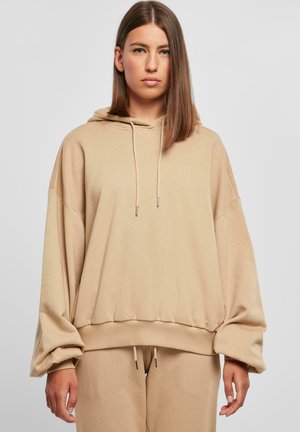 Beige oversized hettegenser med snorhette, ribbede ermer og en myk tekstur. Stoffet fremstår som glatt og litt elastisk.