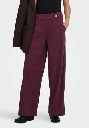 Pantalon large en velours côtelé de couleur bordeaux foncé sur une personne portant un haut noir et tenant une veste marron foncé sur une épaule.