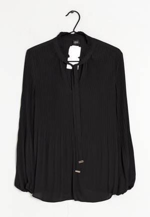 Blouse noire à manches longues en tissu plissé, avec col à volants et détail de lien sur le devant, présentée sur un cintre noir sur fond blanc.