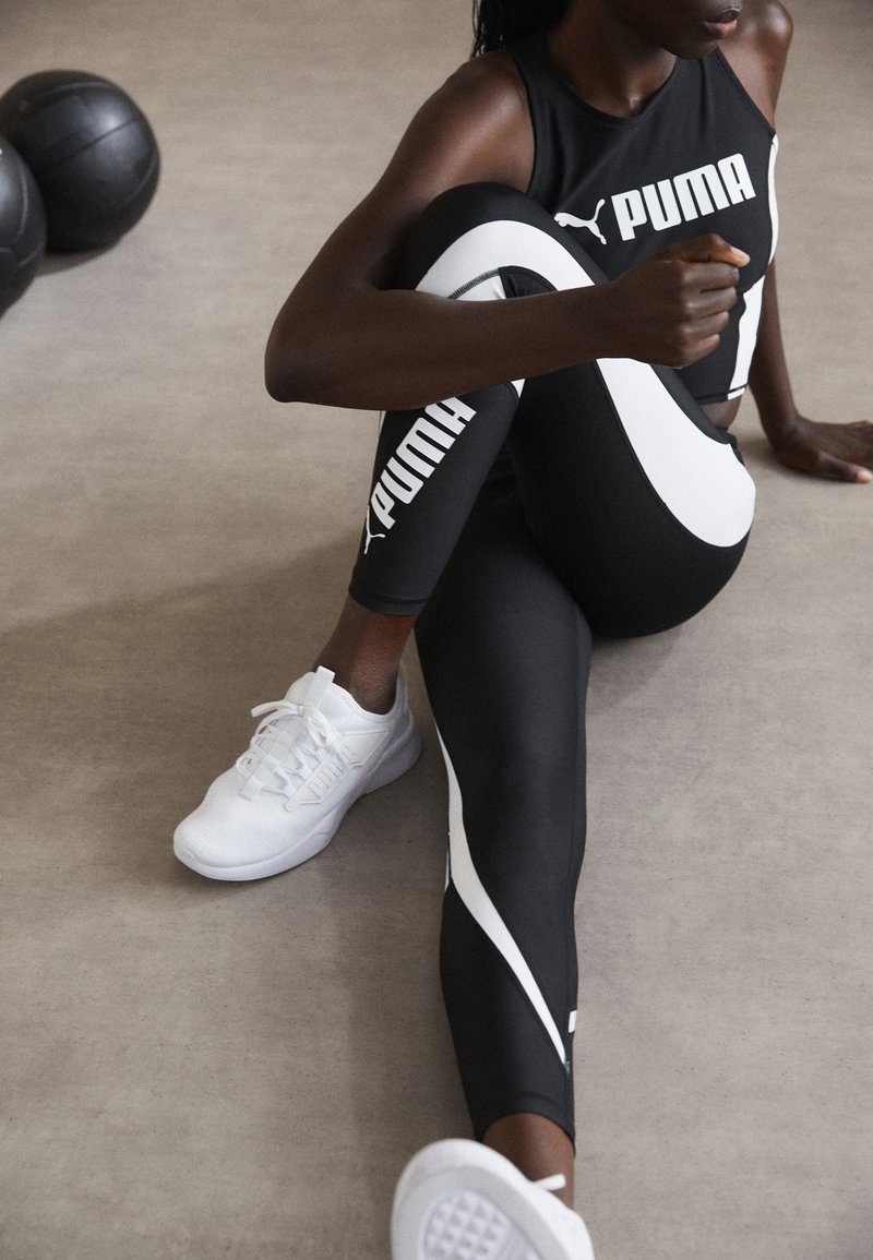 Top atlético negro con logo blanco de Puma, combinado con leggings negros con acentos laterales blancos. Zapatillas de deporte blancas, con textura suave.