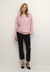 Halvány rózsaszín, túlméretezett pulóver bordázott aljúval, fekete, cropped nadrággal párosítva. A modellen fekete magassarkú cipők vannak. Egyszerű háttér.