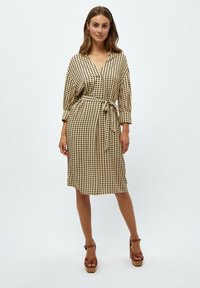 Vyötäröllä oleva, polven mittainen beige mekko mustalla geometrisellä kuviolla, jossa on V-pääntie, pitkät hihat ja löysä istuvuus. Ruskeat platform-korkokengät täydentävät asun.