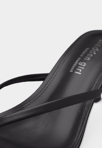 Κοντινή λήψη ενός μαύρου σανδαλιού με λεπτές λουρίδες και ελαφρώς ανυψωμένη φτέρνα σε λευκό φόντο.