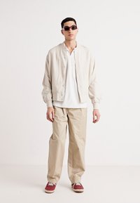 Veste bomber beige clair avec boutons-pression, t-shirt blanc à col en V, pantalons beiges amples et sneakers bordeaux. Des lunettes de soleil sombres complètent la tenue.
