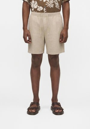 Mies, jolla on päällä beiget nauhakiristimellä varustetut pellavashortsit ja ruskeat kaksinkertaiset remmilenkkarit, seisoo yksivärisen valkoisen taustan edessä.