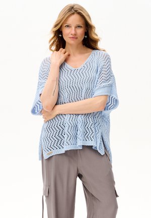 Kobieta o blond włosach, ubrana w jasnoniebieski, ażurowy sweter i luźne, szare spodnie typu cargo, stoi z rękami skrzyżowanymi, z jedną dłonią blisko twarzy.