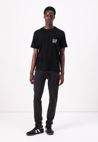 Camiseta de algodón negra de manga corta con pequeño logo blanco de "GAP" en el pecho, combinada con jeans gris oscuro y zapatillas negras.