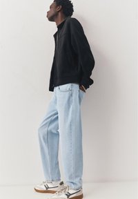 Jonge man met een zwart jack, lichtblauwe spijkerbroek en witte sneakers, staand zijwaarts tegen een witte muur met handen in de zakken.