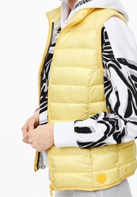 Žlutá péřová vesta s prošívaným designem, vybavená předním zipem a kulatou nášivkou na lemu. Nositá přes černobíle vzorovaný top.