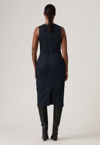 Γυναίκα με πλεξούδες φορώντας ένα μανίκι σκούρο μίντι φόρεμα από τζιν και μαύρες ψηλές μπότες μέχρι το γόνατο με τακούνι, στέκεται με την πλάτη προς την κάμερα.