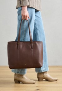 Brun læder taske med side-syninger, to lange håndtag, logo i guld hardware, parret med lyseblå vide ben jeans og beige ankelstøvler.