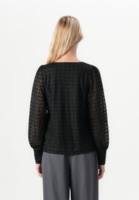 Kvinna med långt blont hår som bär en svart spetsblus med långa ärmar och grå byxor, sedd bakifrån mot en vit bakgrund.