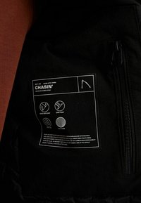 Sort jakke indvendig label med hvide ikoner og tekst, der angiver egenskaber: vandafvisende, vindtæt og termisk isolering. Lynlåsdetalje synlig.