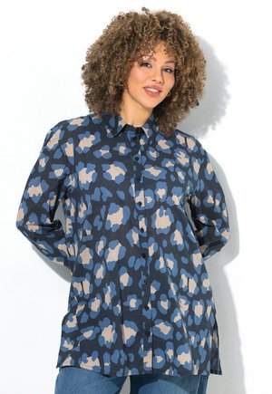 Frau mit lockigem Haar lächelnd, trägt ein langärmliges Hemd mit blau-beigem abstraktem Muster und blaue Jeans.