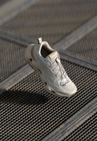 Cream-colored athletic shoe featuring a textured upper with mesh panels and reinforced accents, displayed against a patterned surface.