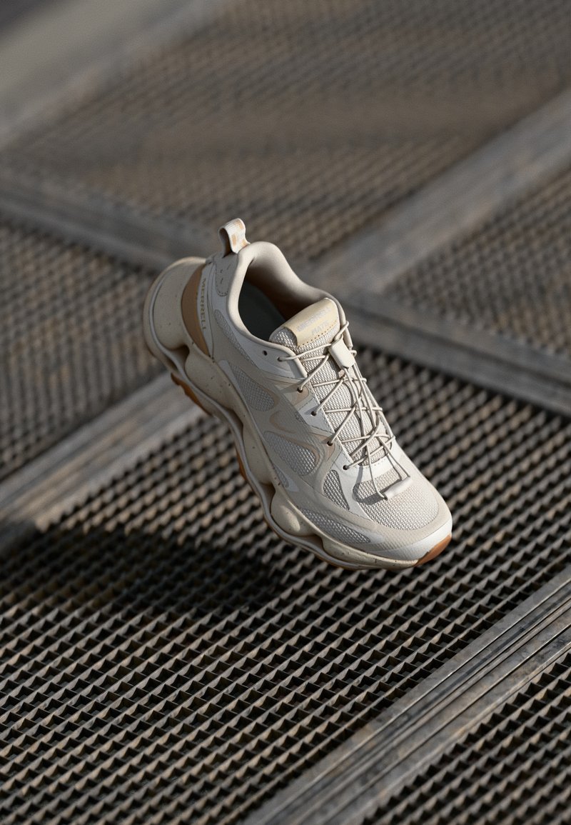 Cream-colored athletic shoe featuring a textured upper with mesh panels and reinforced accents, displayed against a patterned surface.