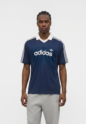 Joven de pie con una camiseta azul marino de Adidas con cuello blanco y rayas, y pantalones deportivos Adidas de color gris claro.