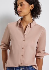 Femme aux cheveux courts et bouclés, portant une chemise beige à manches longues retroussées et un jean bleu, regardant sur le côté.