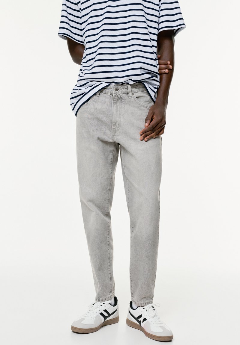 Person in hellblauen Jeans, weißen Sneakers mit schwarzen Streifen und einem navyblauen und weißen gestreiften Kurzarmhemd, steht mit verschränkten Armen hinter dem Rücken.