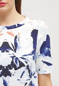 Blusa floreale a maniche corte in bianco, blu e rosa chiaro; presenta un collo rotondo e un motivo di fiori astratti in varie tonalità.