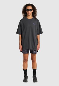 Czarna, oversize koszulka z krótkim rękawem, z fioletowym nadrukiem na klatce piersiowej, zestawiona z wzorzystymi czarnymi szortami i czarnymi skarpetkami.