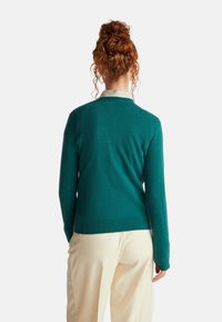 Tyrkysový pletený kardigan s V-výstřihem a dlouhými rukávy, s hladkou texturou a žebrovaným lemem. Nositý přes pruhovanou košili s límečkem.