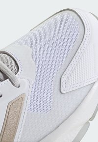 Hvit atletisk sko med mesh-overdel, semsket skinn-detaljer og en teksturert såle. Designet inkluderer en metallisk gulldetalj på siden.