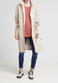Beige, leichter Trenchcoat mit einer Knopfleiste vorne, seitlichen Taschen und hochgerollten Ärmeln, kombiniert mit einem rostfarbenen Oberteil und blauen Jeans.