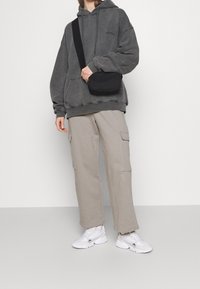 Persona che indossa un hoodie grigio scuro oversize, pantaloni cargo beige, sneakers bianche e una borsa a tracolla nera su uno sfondo neutro.