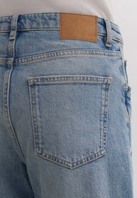 Jeans de mezclilla azul claro con un parche de cuero marrón en la cintura y dos bolsillos traseros con costura visible. Tela texturizada y casual.