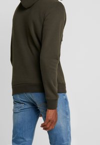 Felpa verde oliva con finitura texturizzata, dotata di cappuccio e polsini a coste, abbinata a jeans in denim blu con dettagli di cucitura visibili.