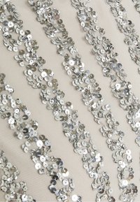 Tissu beige orné de sequins argentés disposés en bandes verticales, présentant des tailles variées et des surfaces réfléchissantes pour un aspect texturé.