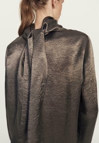 Bluzka z długim rękawem, z metalicznego brązowego materiału o zmarszczonej fakturze, z dużą kokardą na plecach przy szyi.