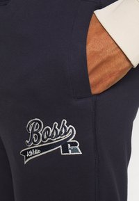 Hand i fickan på mörkblå byxor med broderad "Boss Athletic"-logotyp i vit och marinblå tråd, närbild.