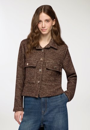 Giacca marrone strutturata con colletto, due tasche sul petto e bottoni argentati, abbinata a jeans blu a vita alta.