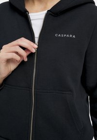 Musta vetoketjuhuppari, joka on valmistettu pehmeästä kankaasta. Hupparissa on etutasku, hopeinen vetoketju ja valkoinen "CASPARA"-logo vasemmassa rinnassa.