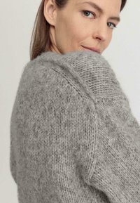 Suéter gris de punto con una textura suave, que presenta un escote ancho y un destacado detalle de costura en los hombros.