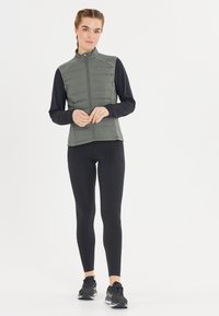Vadderad väst i olivgrön färg med hög krage, dragkedja fram och en stilren design, kombinerad med en svart långärmad topp och svarta leggings.