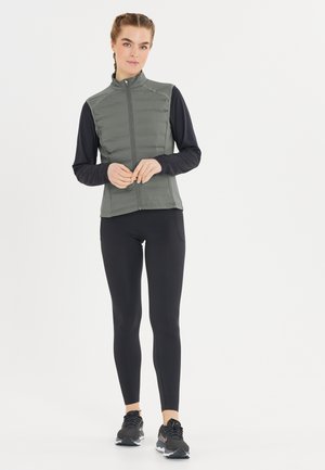 Gepolsterte olivgrüne Weste mit hohem Kragen, Reißverschluss vorne und schlankem Design, kombiniert mit einem schwarzen Langarmoberteil und schwarzen Leggings.