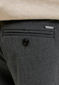 Πίσω τσέπη σκούρου γκρι παντελονιού με κουμπί και μικρό μεταλλικό ταμπελάκι με την επιγραφή Jacks Jones, κάτω από μπεζ πλεκτό πουλόβερ.