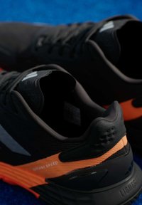 Μαύρα αθλητικά παπούτσια με πορτοκαλί λεπτομέρεια, με υφή στην άνω πλευρά, ελαφρύ σχεδιασμό και λογότυπο στο πλάι.