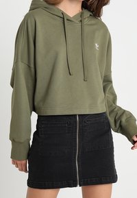 Olive green cropped hoodie with a drawstring, paired with a black denim mini skirt featuring a front zipper and back pockets.