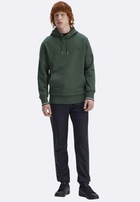 Felpa verde scuro con cappuccio, tasca a marsupio, coulisse regolabile e polsini a costine, abbinata a pantaloni scuri e scarpe nere.